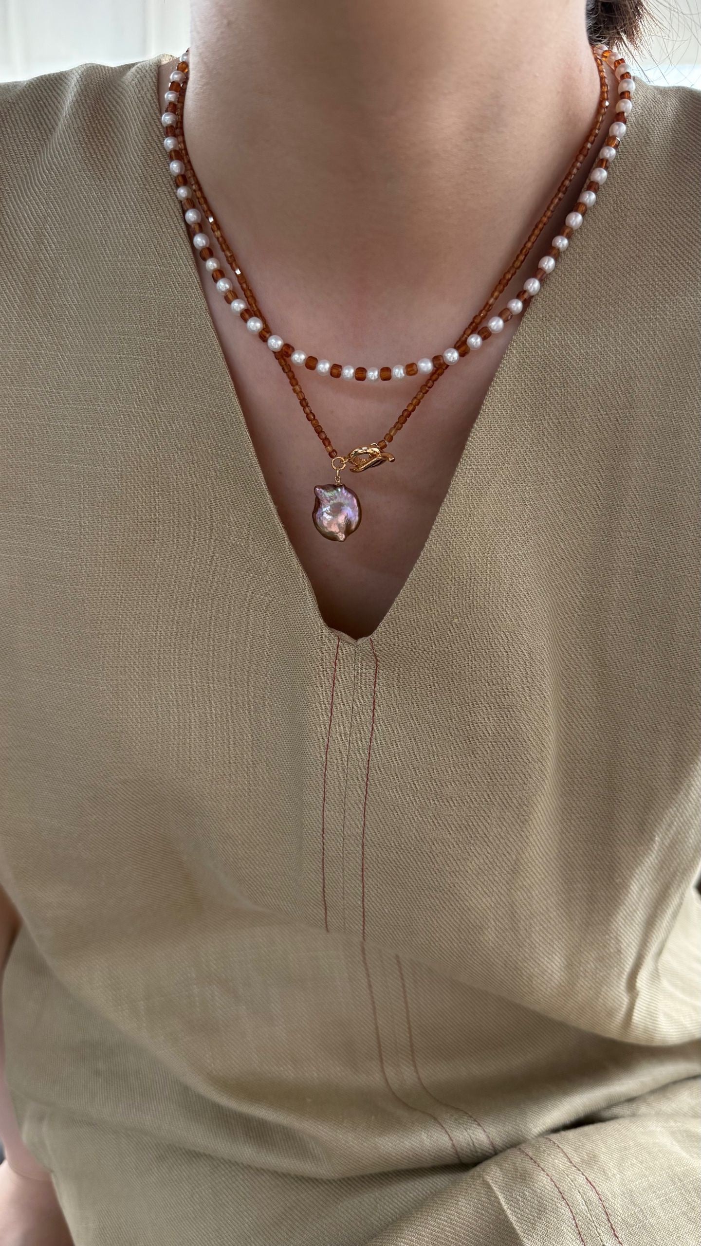 Close-up of a person wearing a pearl necklace with a pendant on a beige top.