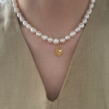 Pearl necklace with a gold pendant worn by a person in a beige outfit.