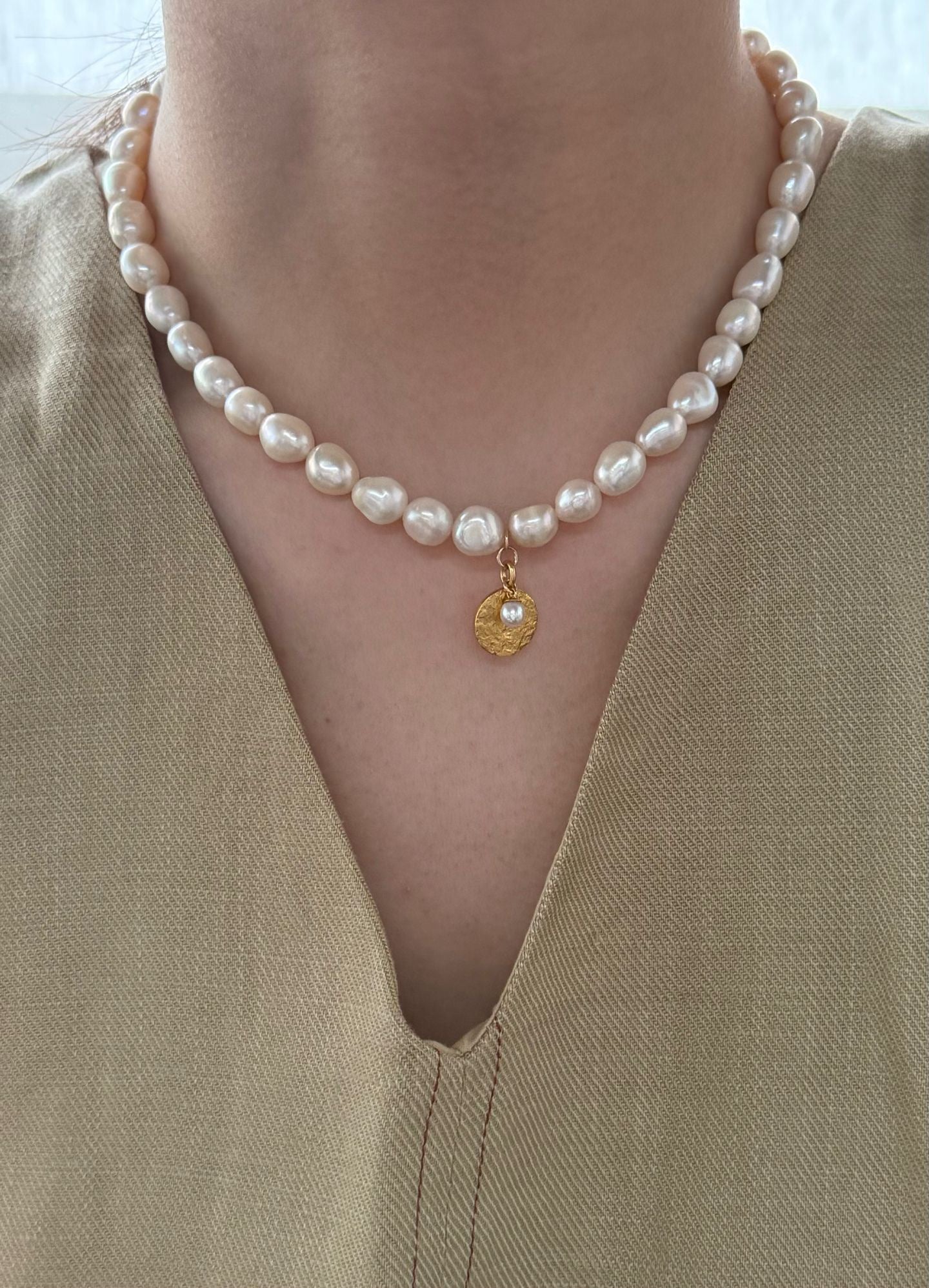 Pearl necklace with a gold pendant worn by a person in a beige outfit.
