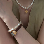 Pearl bracelet with gold heart charm worn on wrist, pearl necklace on neck, against a neutral background
