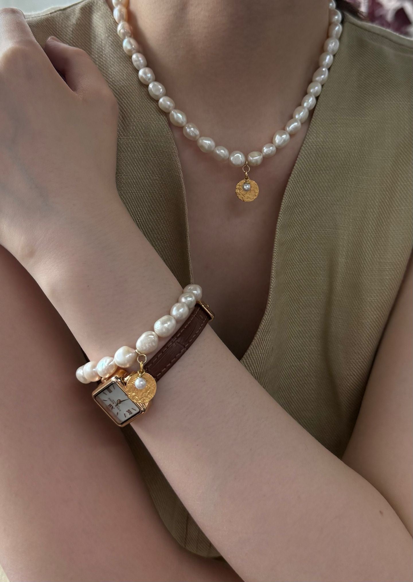 Pearl bracelet with gold heart charm worn on wrist, pearl necklace on neck, against a neutral background
