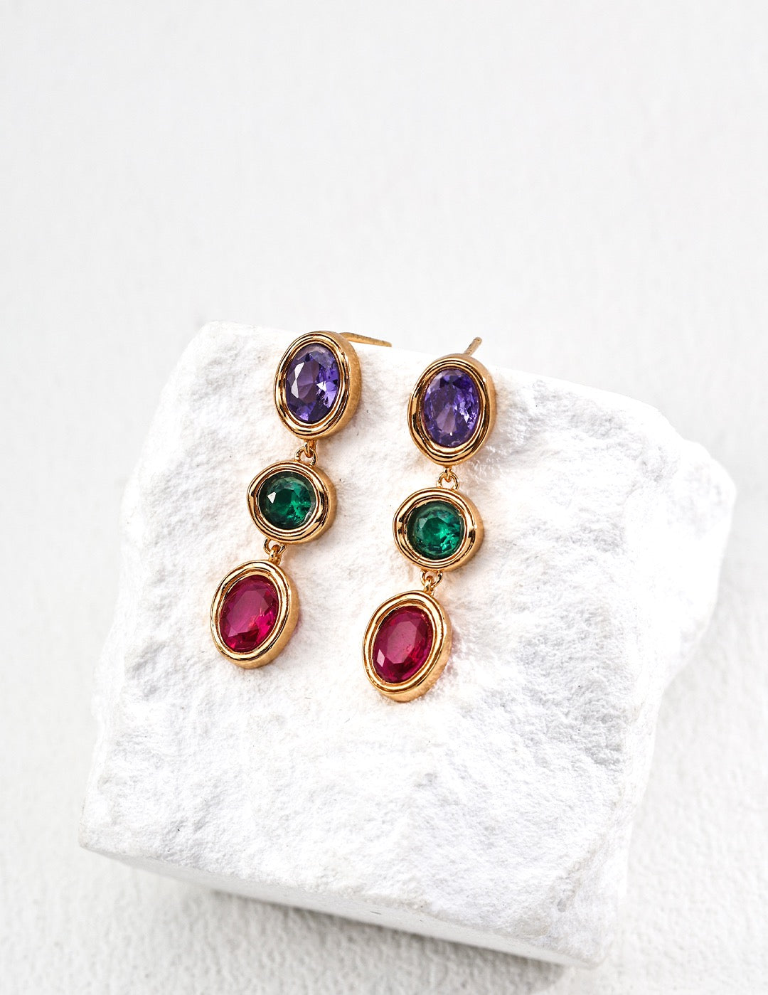 Gold earrings with multicolored gemstones on a white textured surface