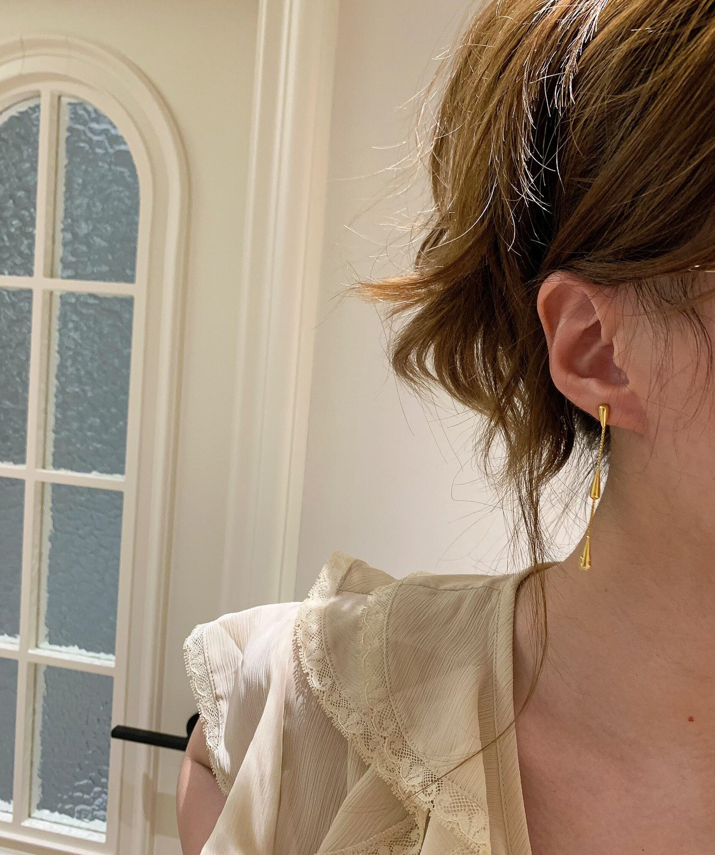 Person wearing a light-colored top with lace details, standing in a room with a window.