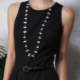 Person wearing a black dress with a long necklace, standing against a neutral background.
