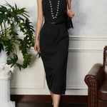 Woman wearing a black dress with a belt, standing indoors next to a plant and a couch.