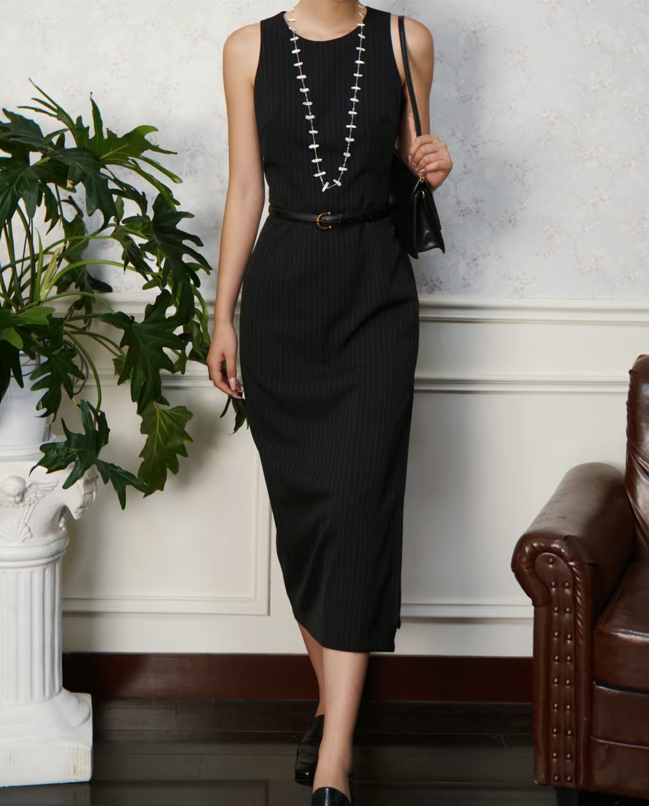 Woman wearing a black dress with a belt, standing indoors next to a plant and a couch.