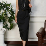 Woman wearing a black dress with a belt, standing indoors next to a plant and a couch.